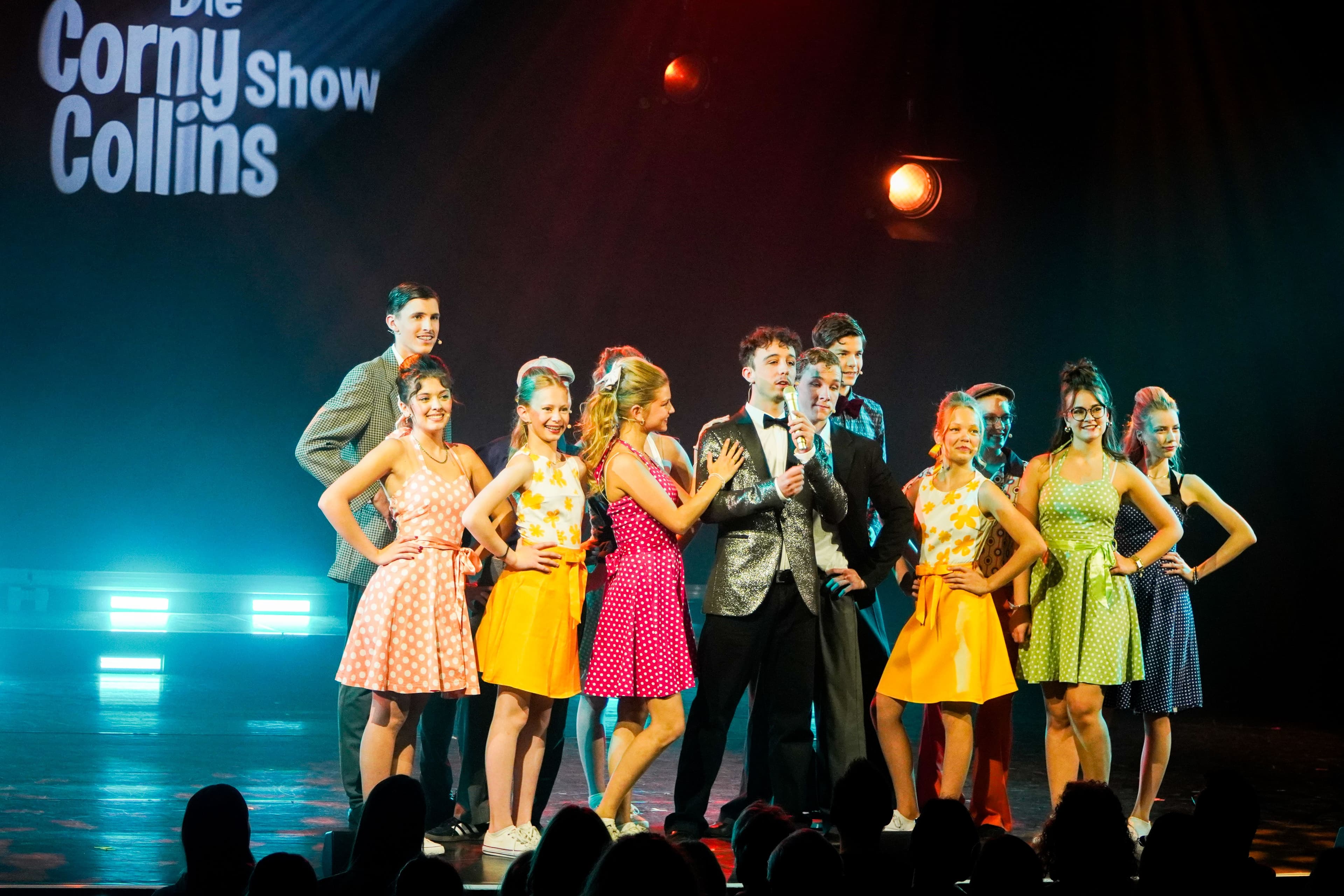 Schauspieler in bunten Kostümen auf einer Bühne, stehen um einen Mann mit Anzug herum.