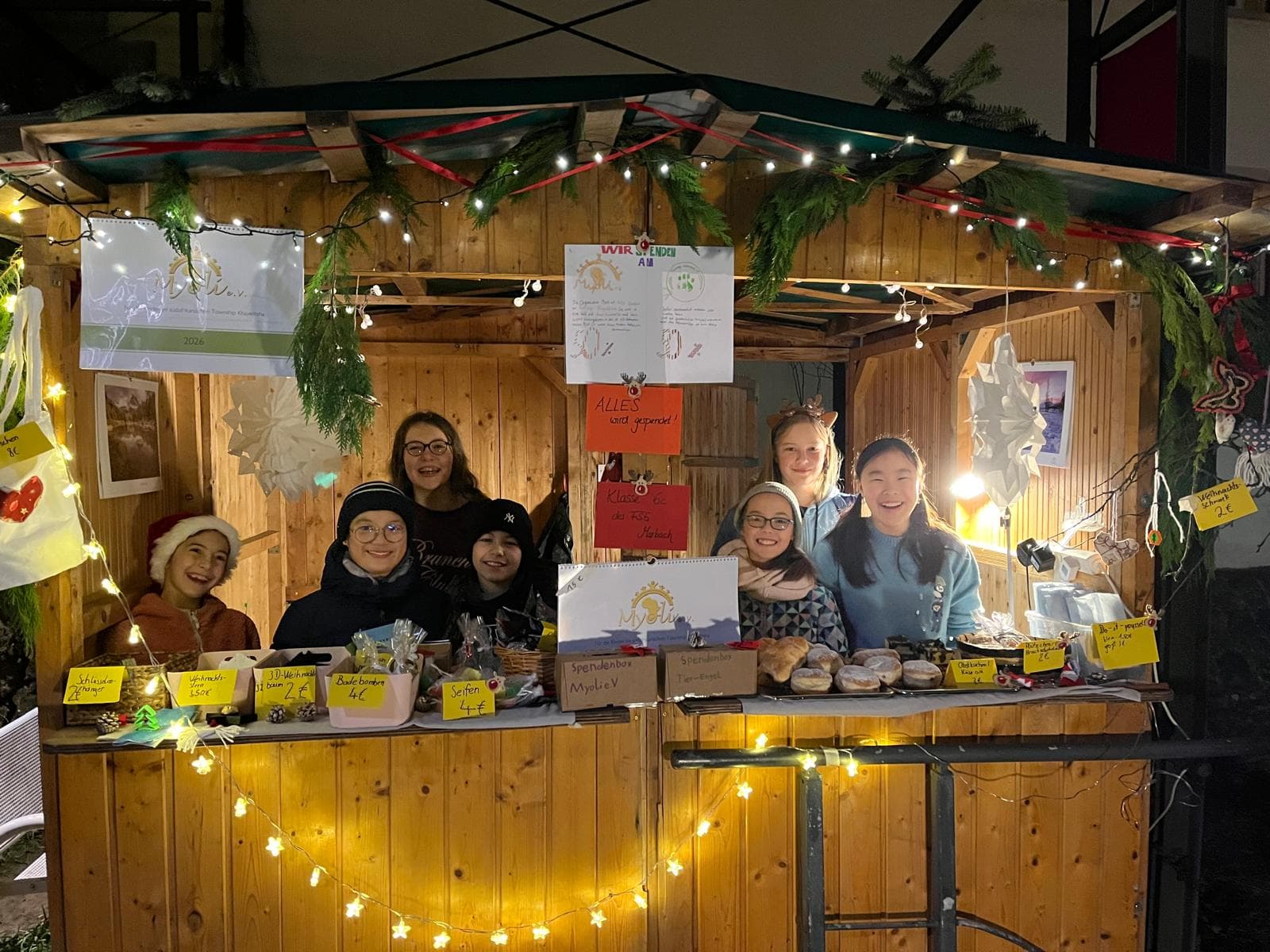A group of smiling children and young adults behind a festive wooden market stall with lights and greenery, selling various items.