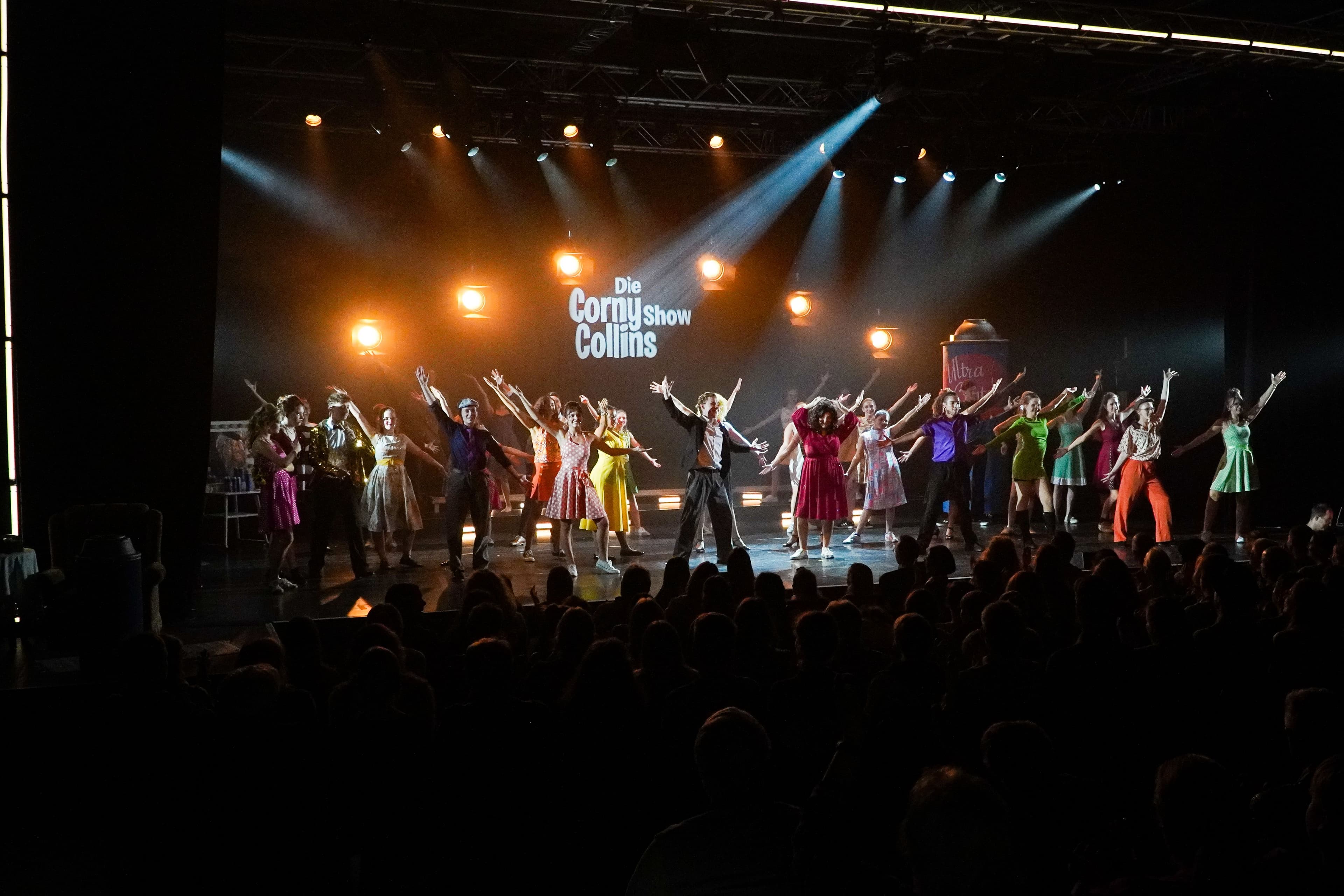 Großes Ensemble in Tanzpose auf einer beleuchteten Bühne, davor Publikum.