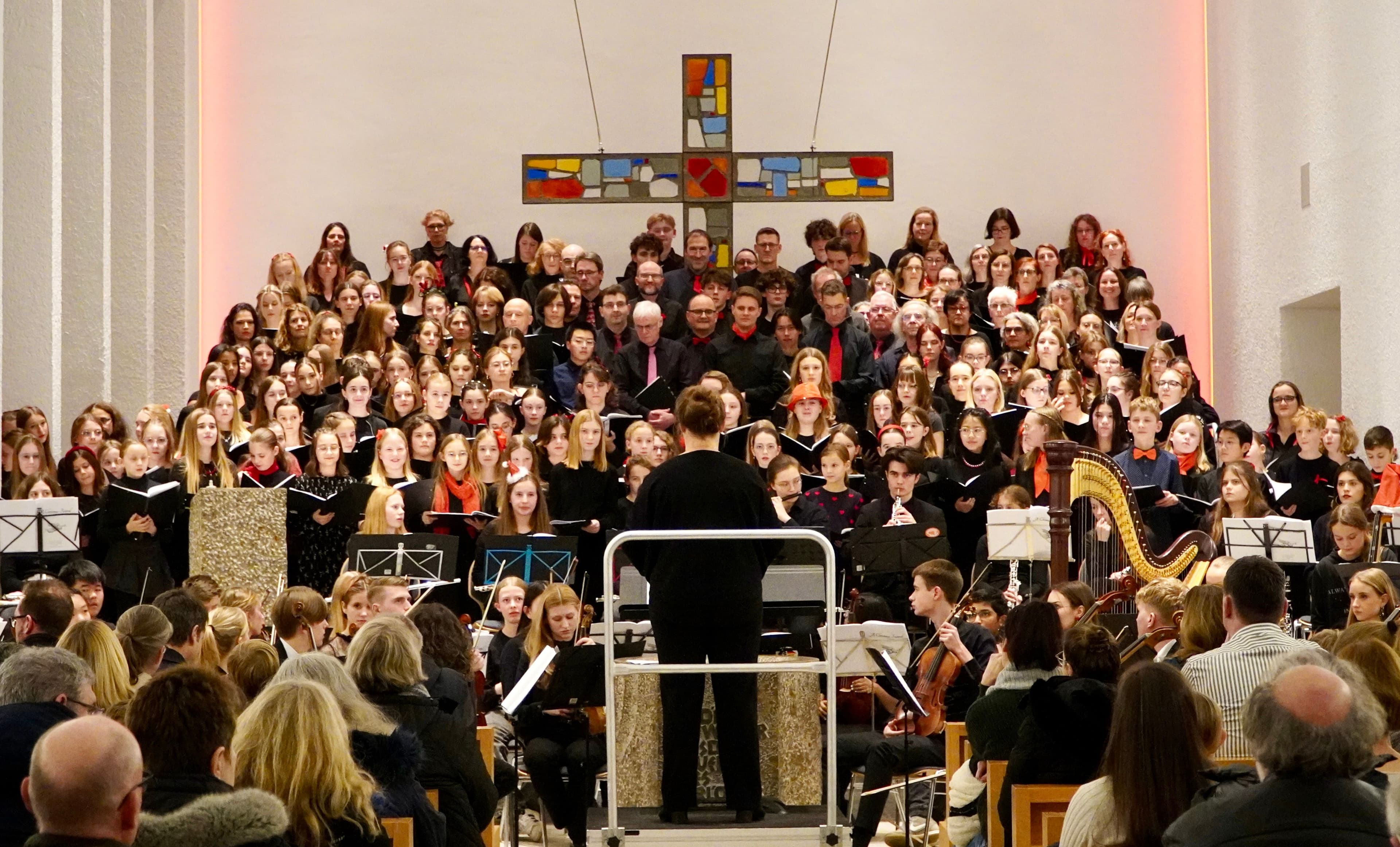 A conductor leads a large choir and orchestra performing in a church, with an audience in the foreground.