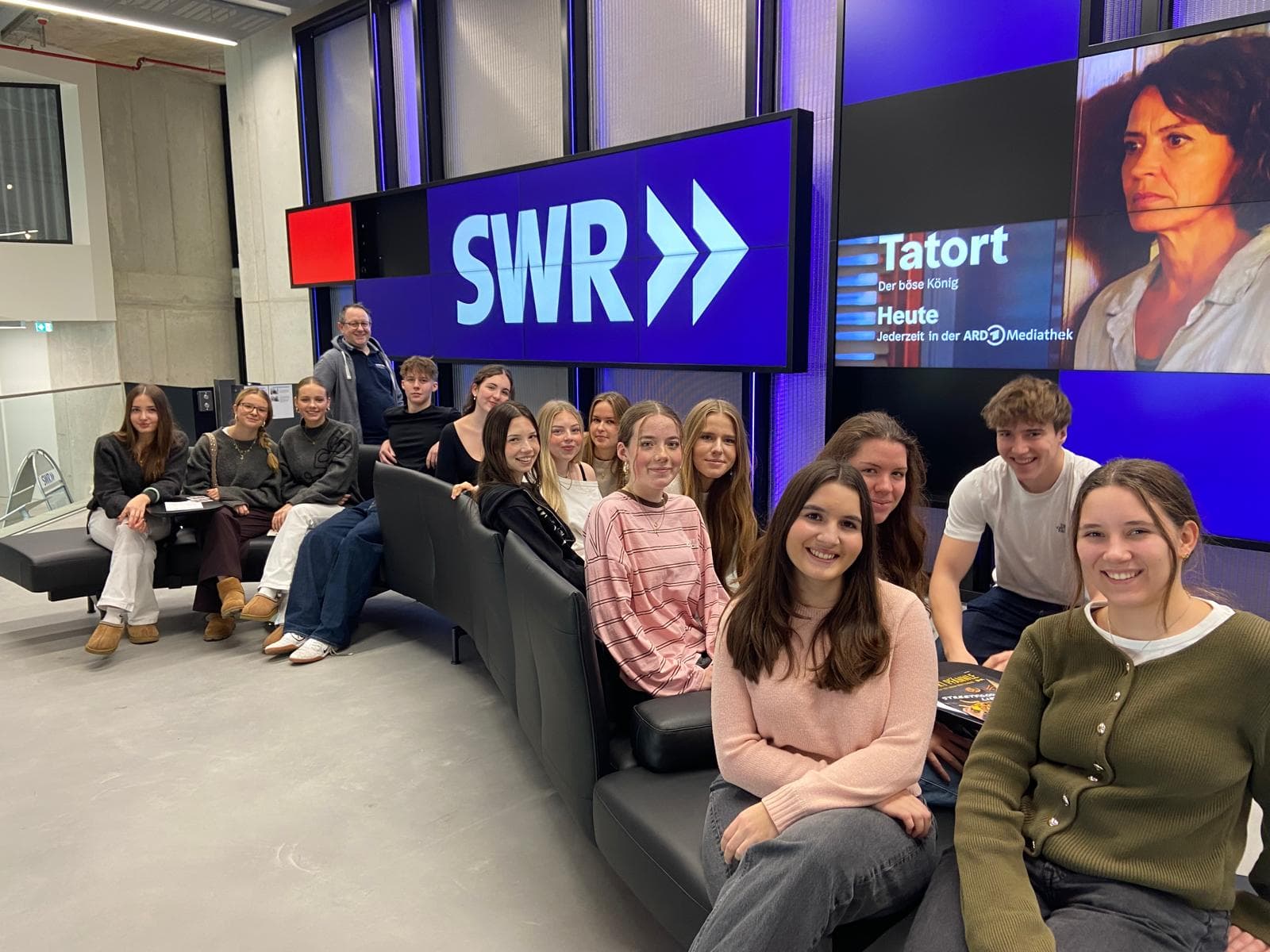 A group of smiling young people and an adult man sit on sofas in a modern media room with large SWR and Tatort screens in the background.