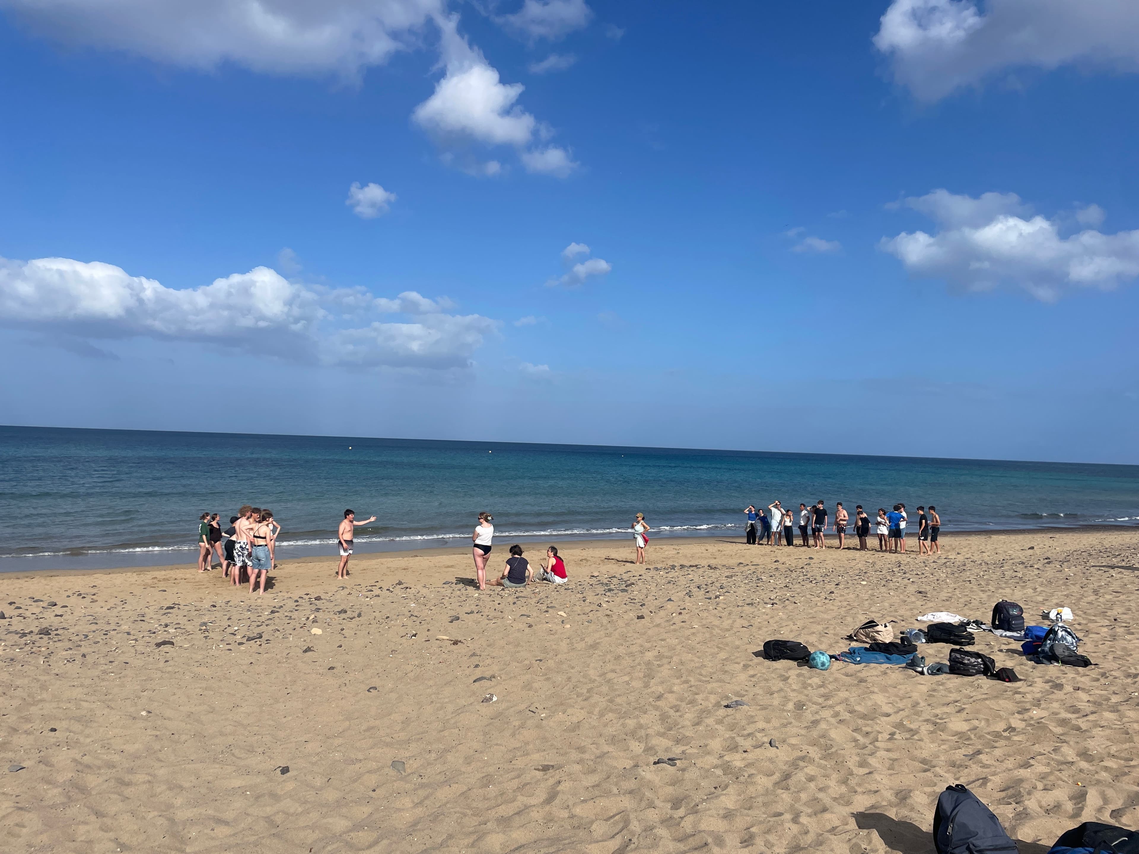 Groups of people gathered on a sandy beach under a blue sky, with the ocean in the background and bags scattered in the foreground.