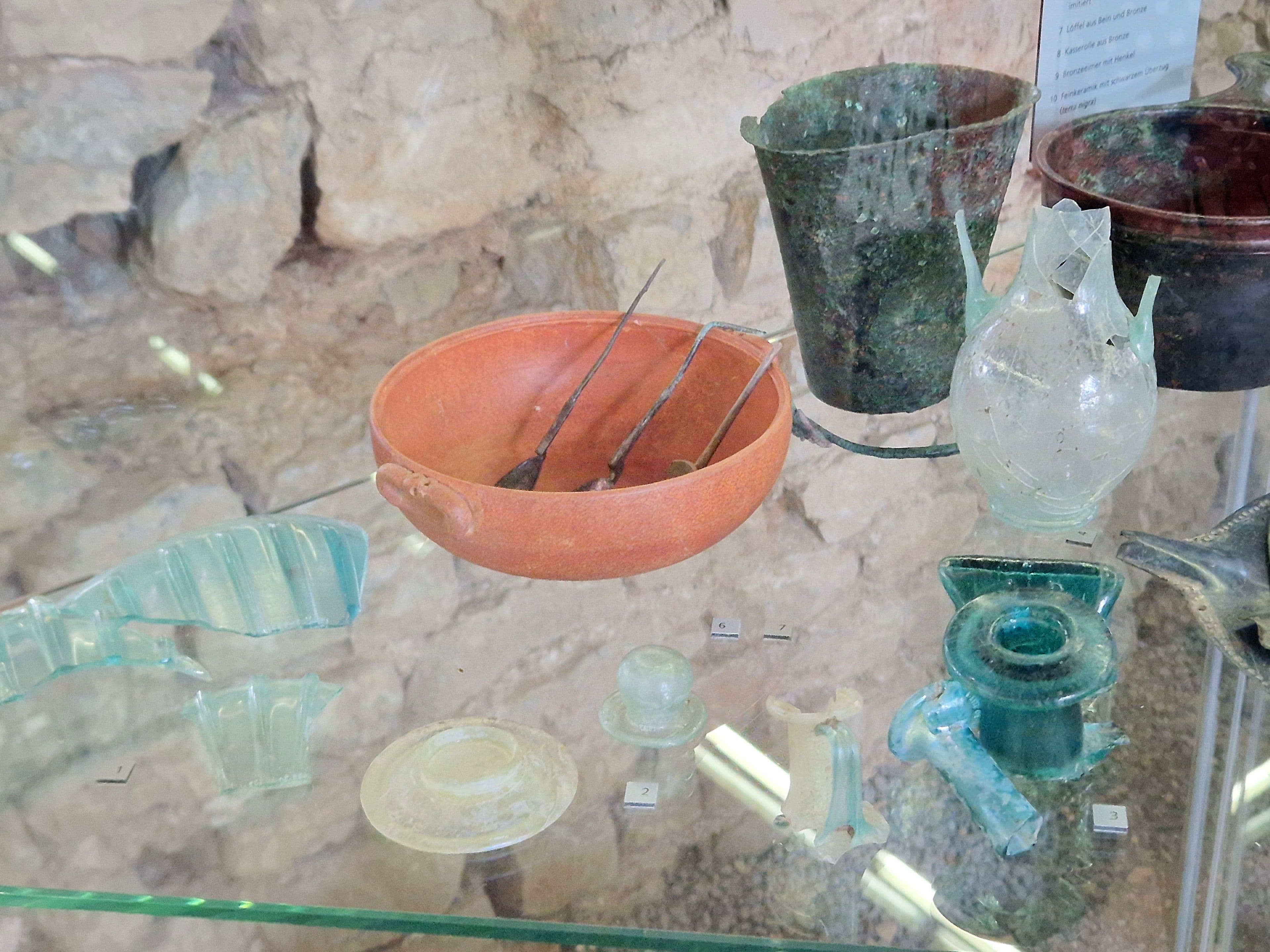 Assortment of ancient artifacts: a terracotta bowl with tools, weathered metal buckets, and various clear and blue-green glass pieces, displayed on a glass shelf.