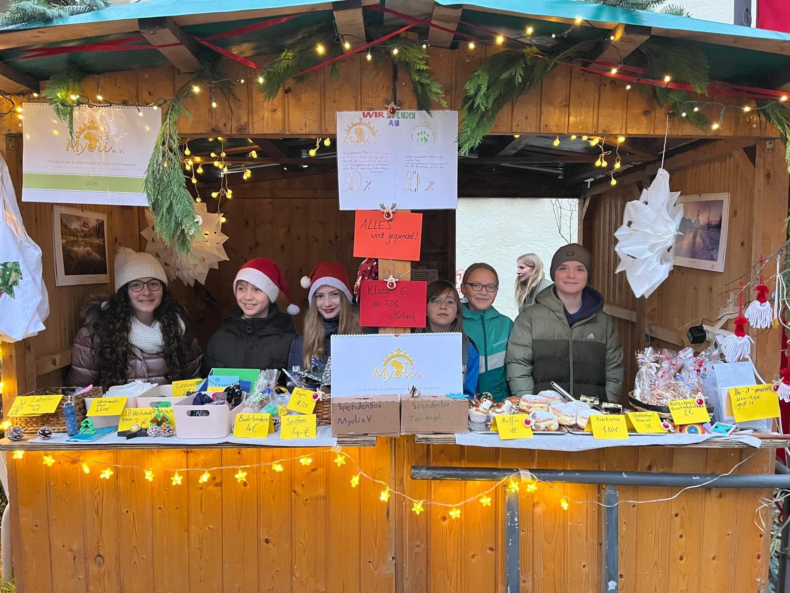 Six children smile from behind a decorated wooden stall at a Christmas market.