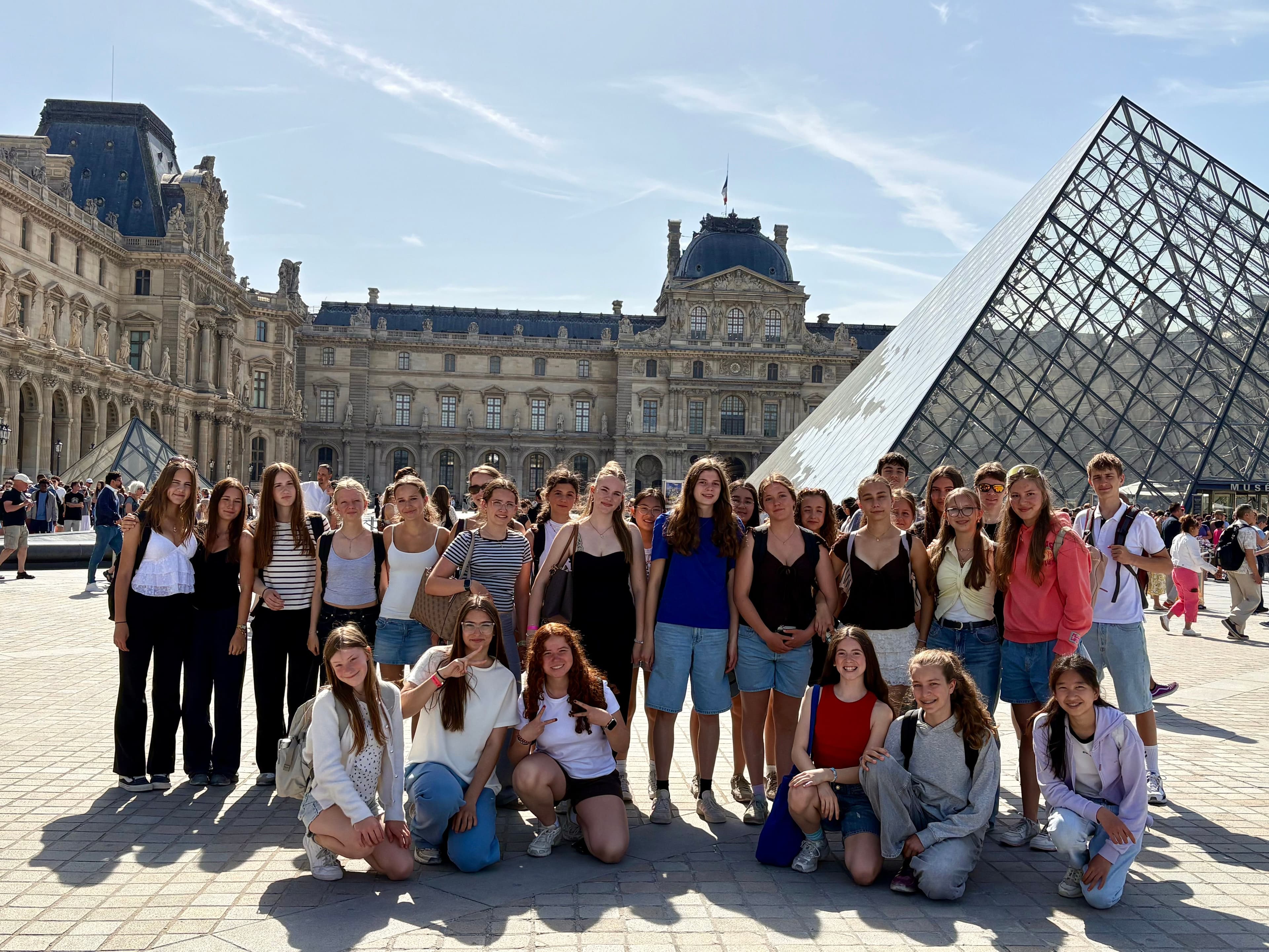 A group of young people posing in front of the Louvre Museum and its glass pyramid in Paris.