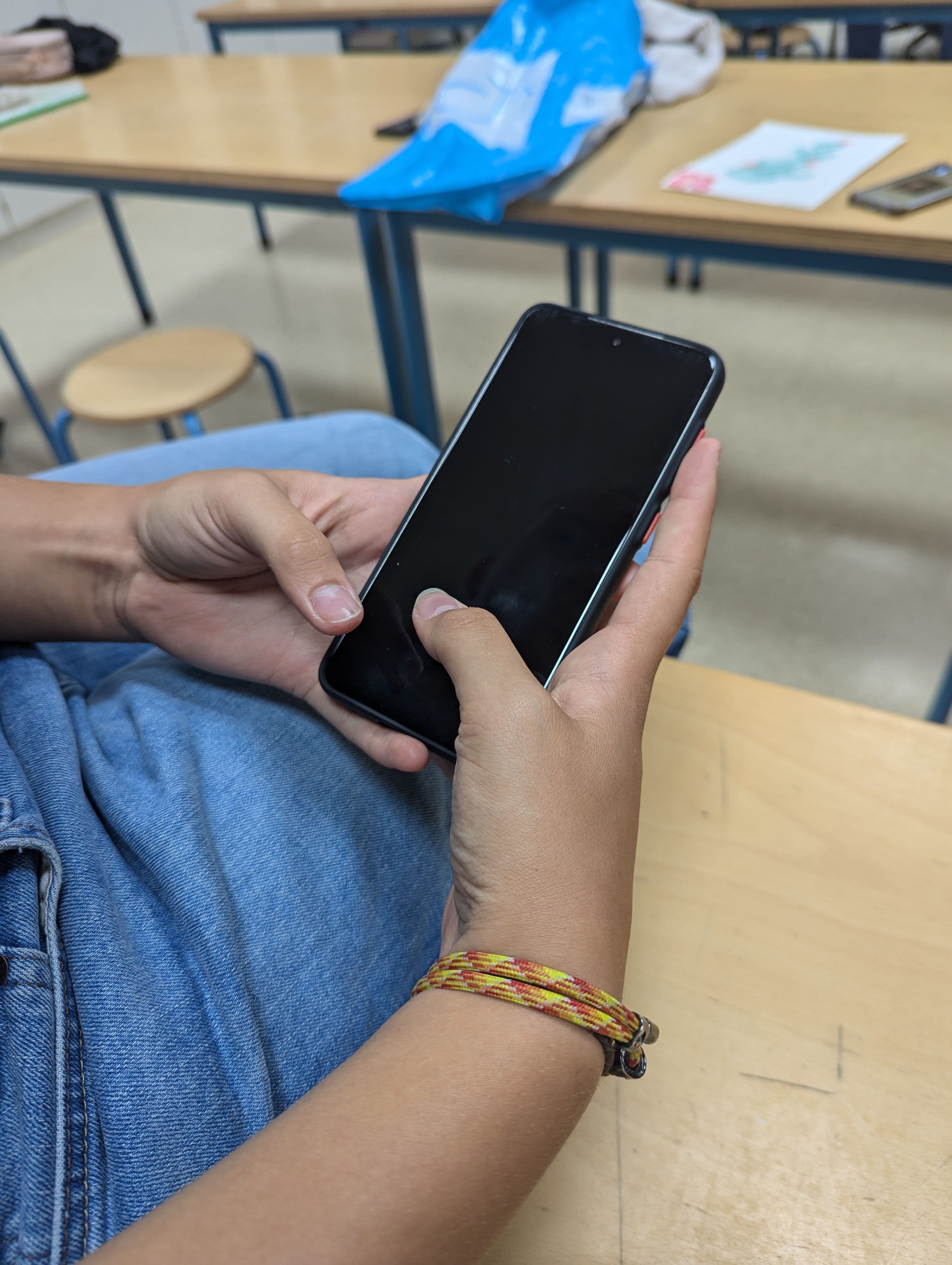 A person wearing blue jeans and a colorful bracelet holds a smartphone with a black screen.