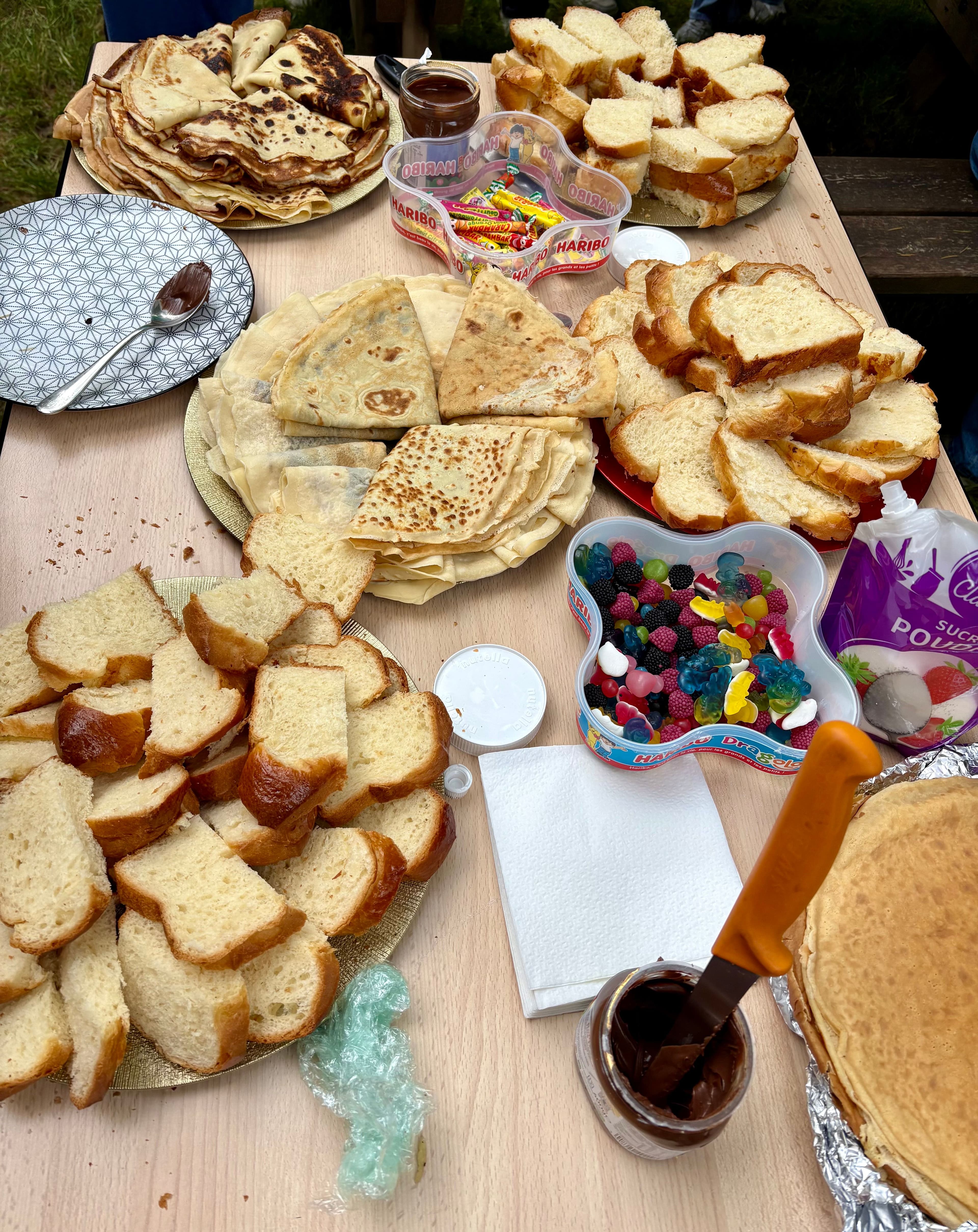 A wooden table overflowing with plates of pancakes, sliced sweet bread, chocolate spread, and colorful candies.