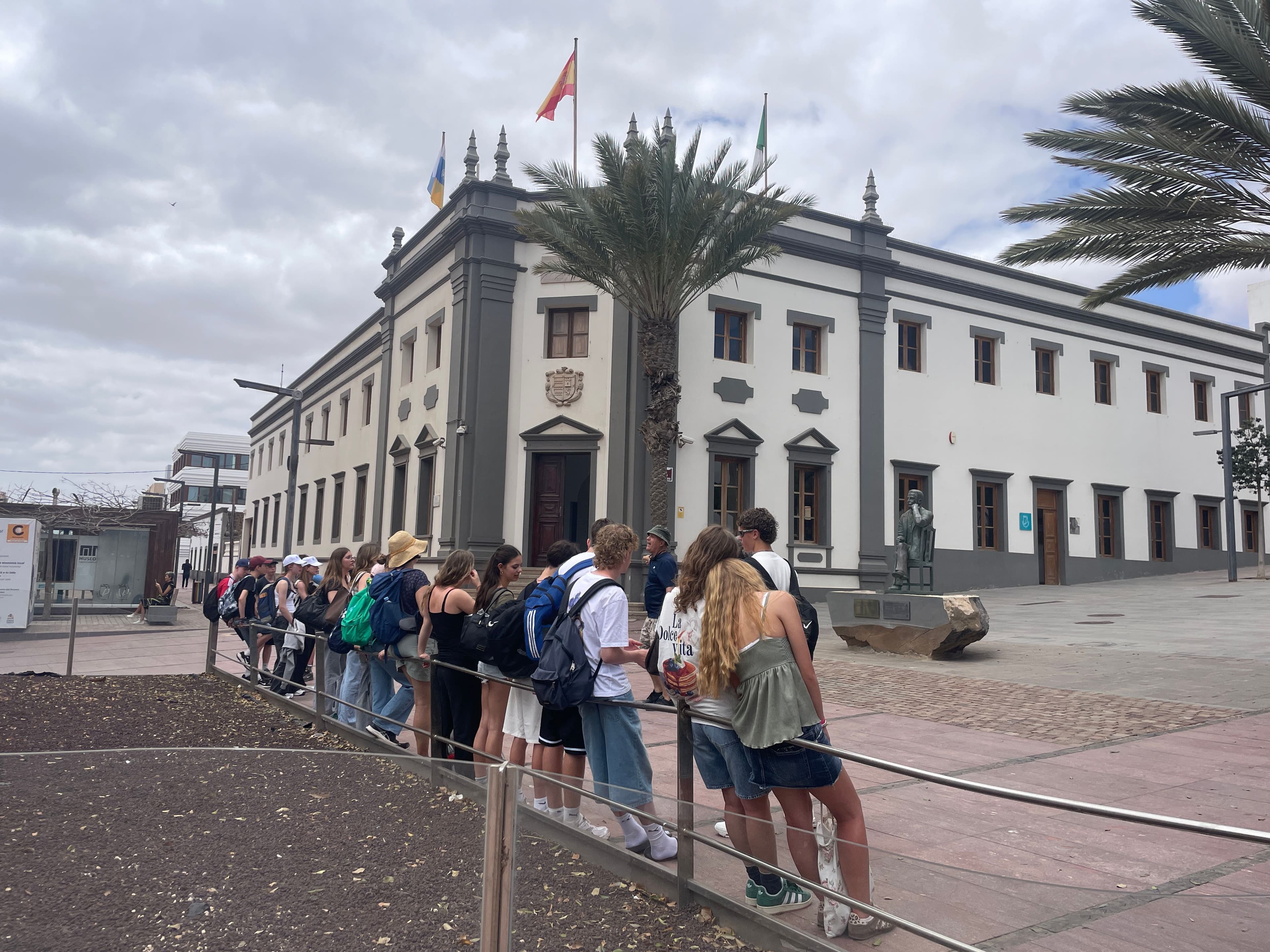 A group of young people queues along a railing in front of a white building with gray trim, a Spanish flag, and palm trees under a cloudy sky.