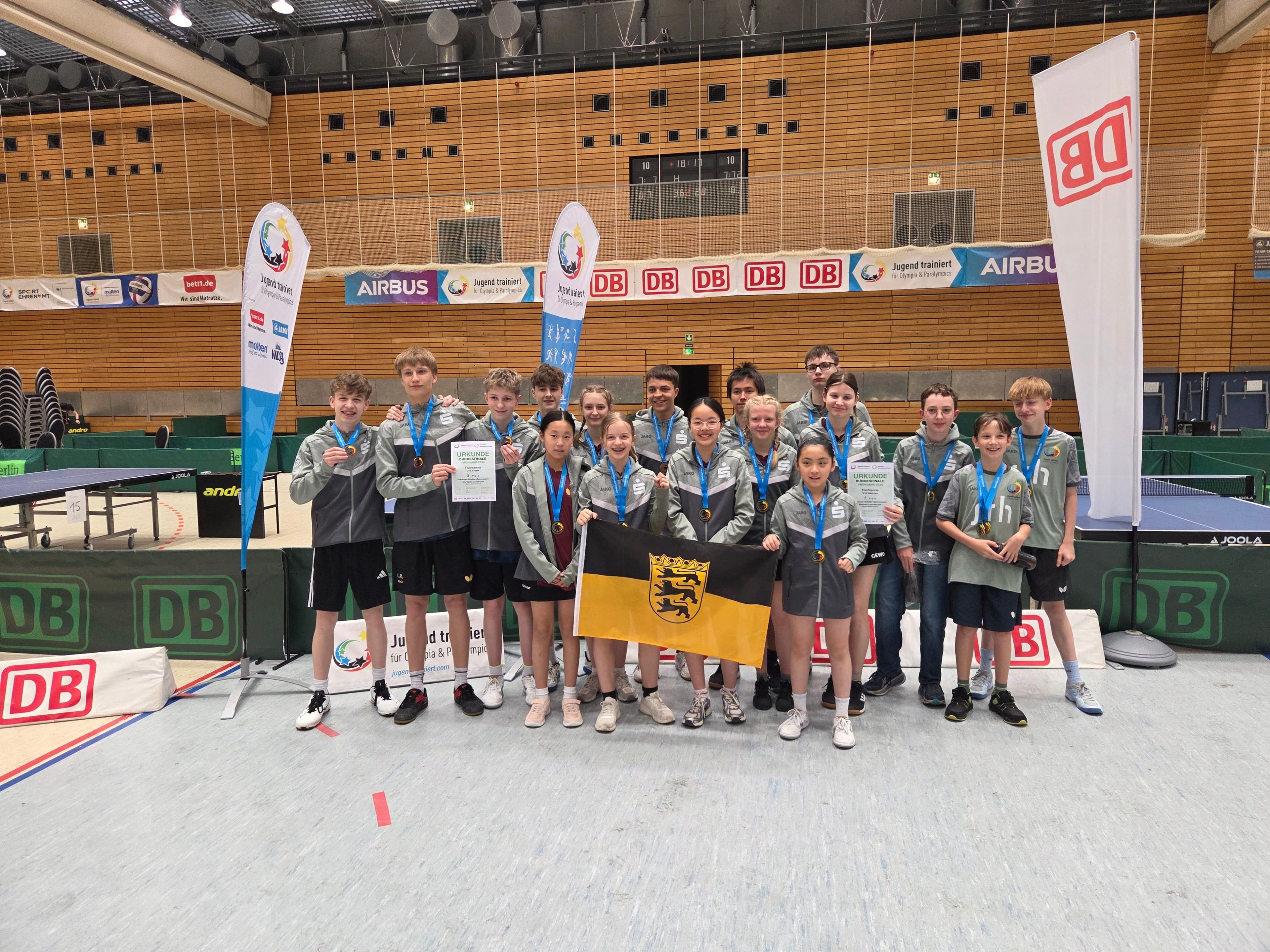 A group of young athletes wearing medals and holding certificates, with a Baden-Württemberg flag, in a sports hall.