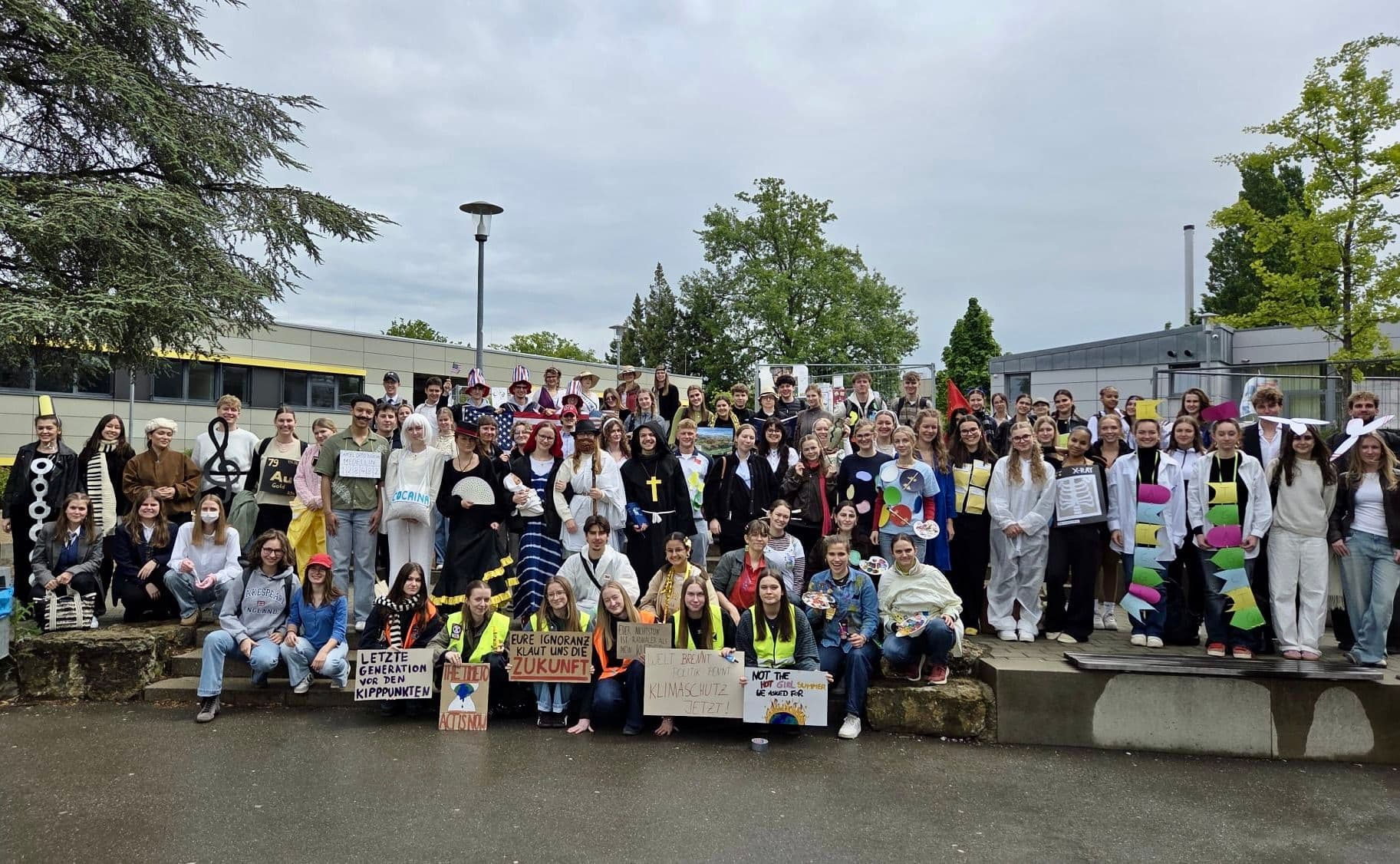 A large group of students in various costumes holding signs addressing social and environmental issues, gathered outdoors.