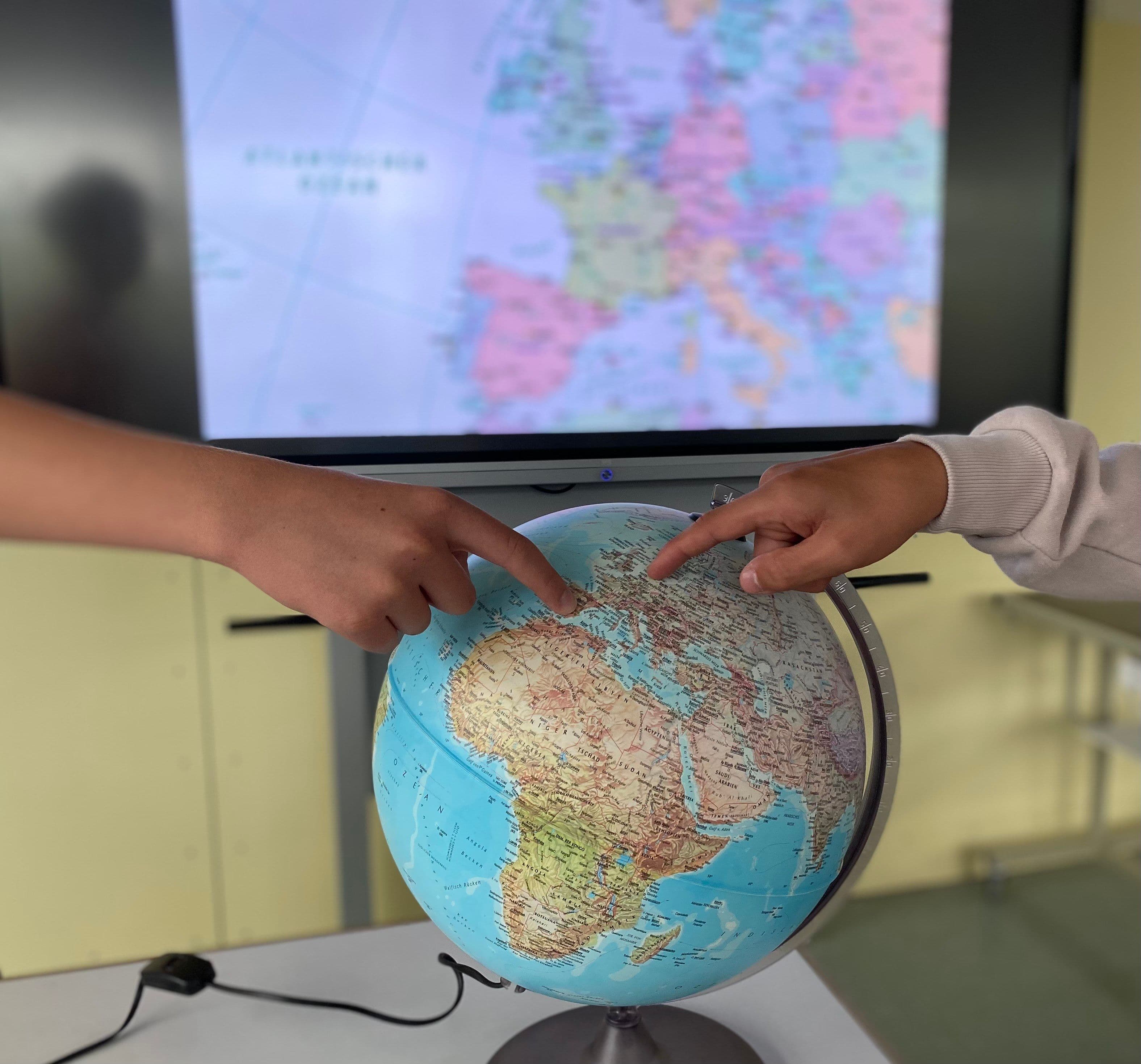 zei Hände zeigen auf einen Globus, der vor einem Bildschirm mit Landkarte von Europa steht