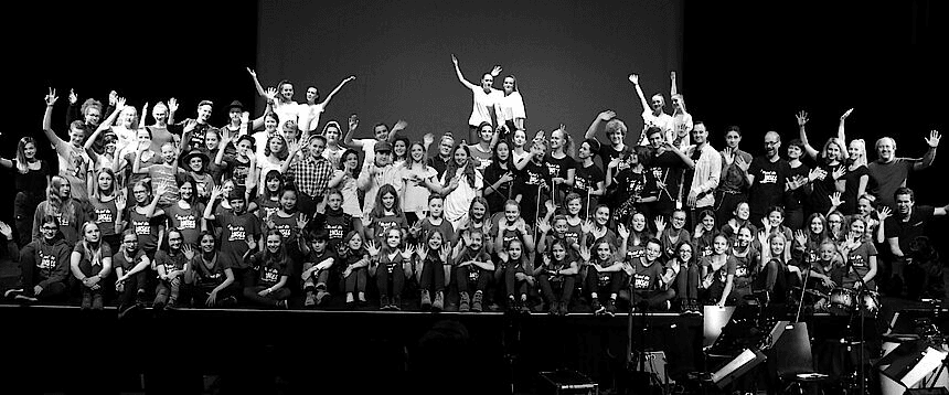 Eine große Gruppe Menschen stehen auf einer Bühne und winken in schwarz-weiß