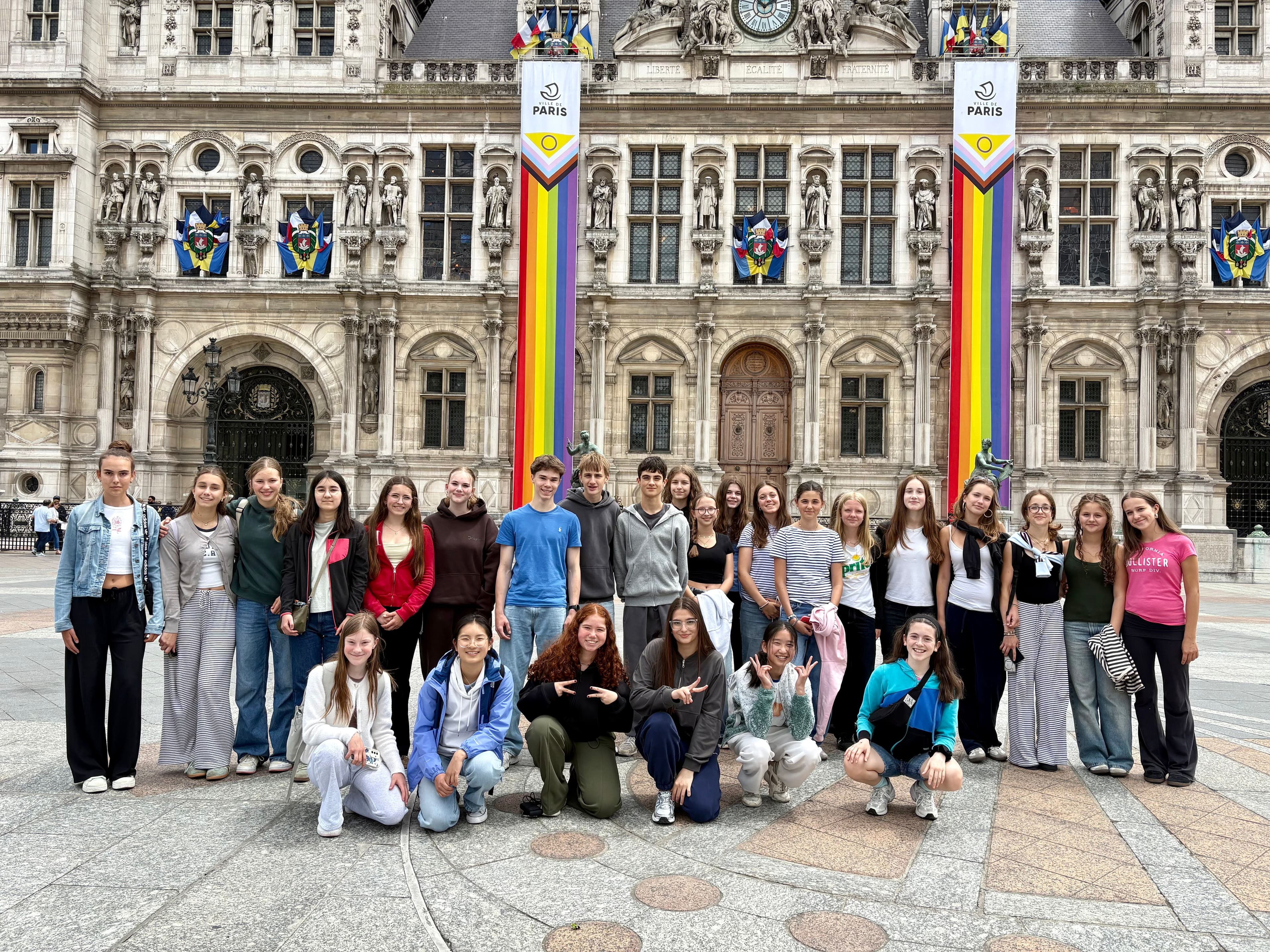 A group of young people posing in front of a grand building adorned with rainbow Paris 2024 Olympic banners.