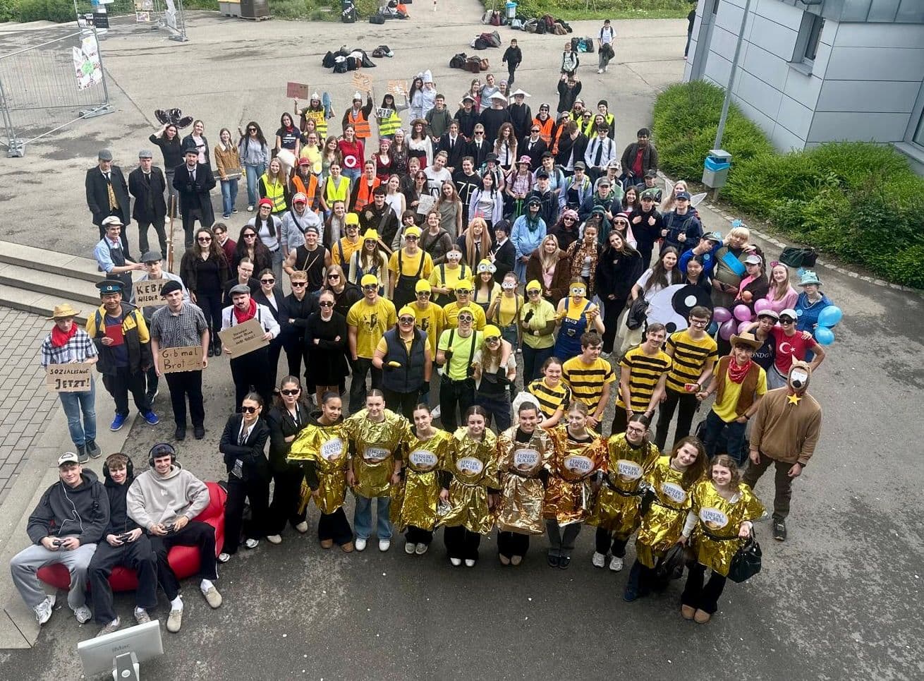 A large group of people in diverse themed costumes, including golden wrappers, bees, and construction workers, pose outdoors.