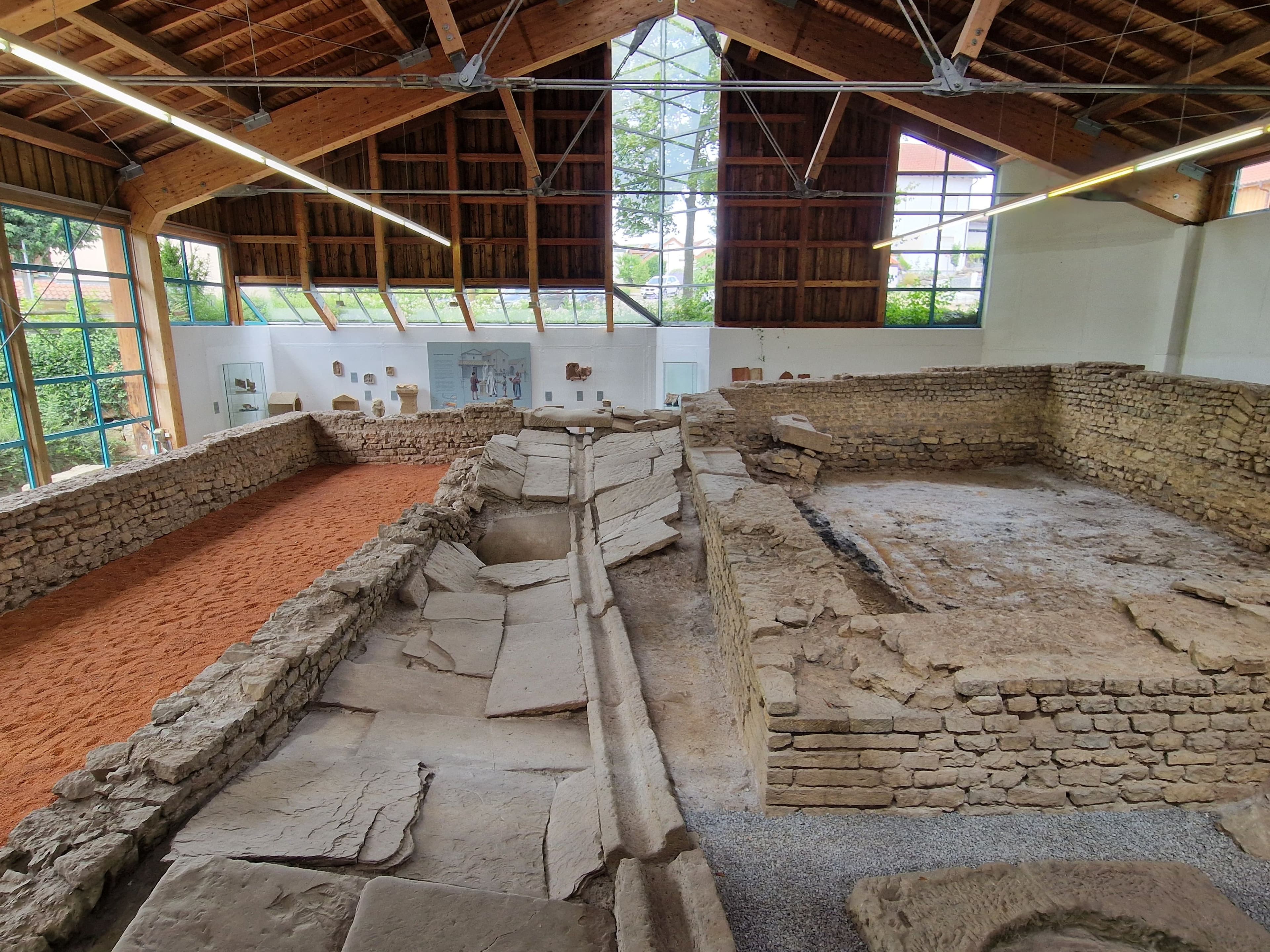 Excavated ancient stone ruins displayed indoors under a modern wooden-beamed roof with large windows.