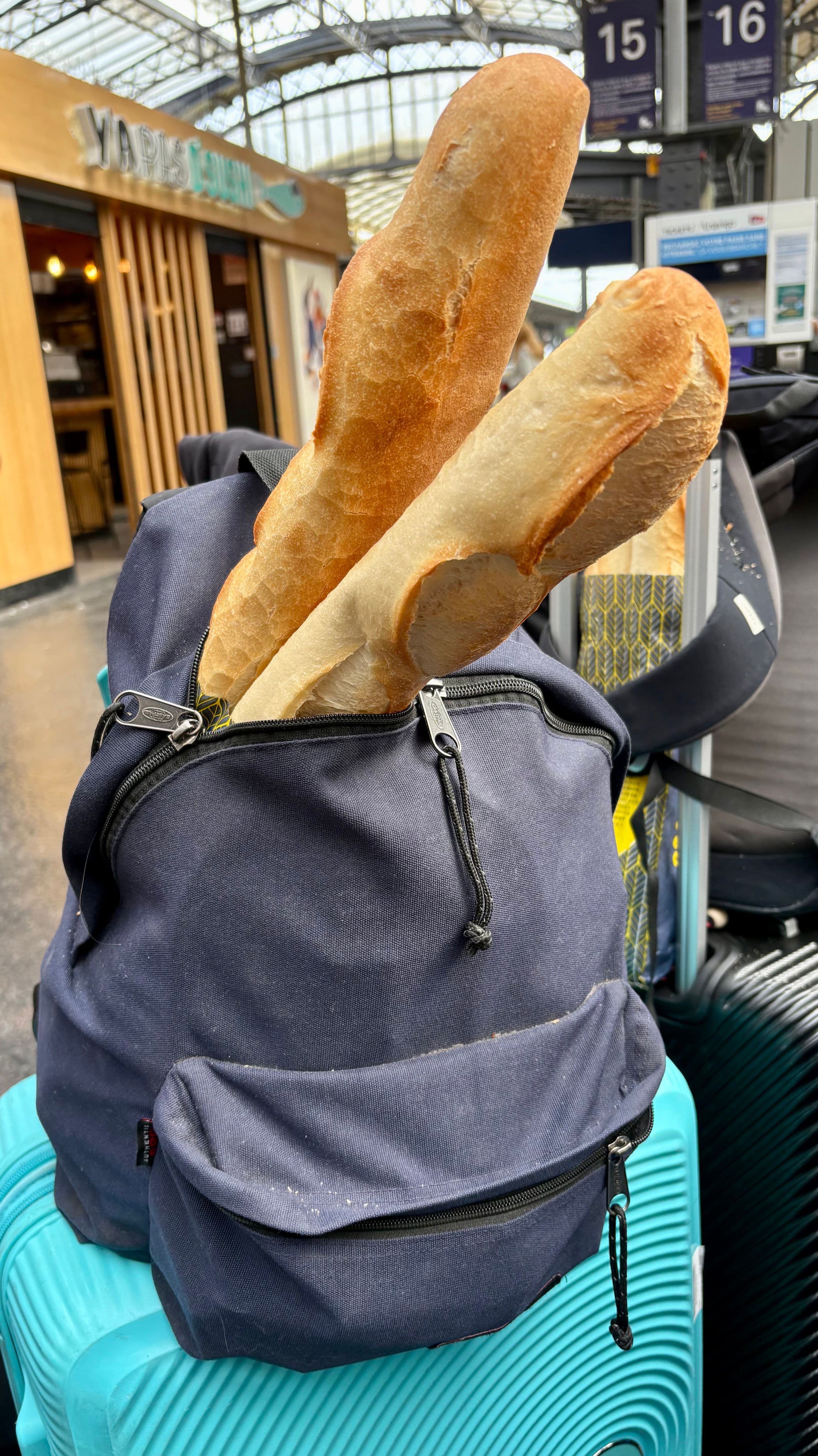 Two baguettes sticking out of a navy blue backpack, which is resting on a teal suitcase in a train station.